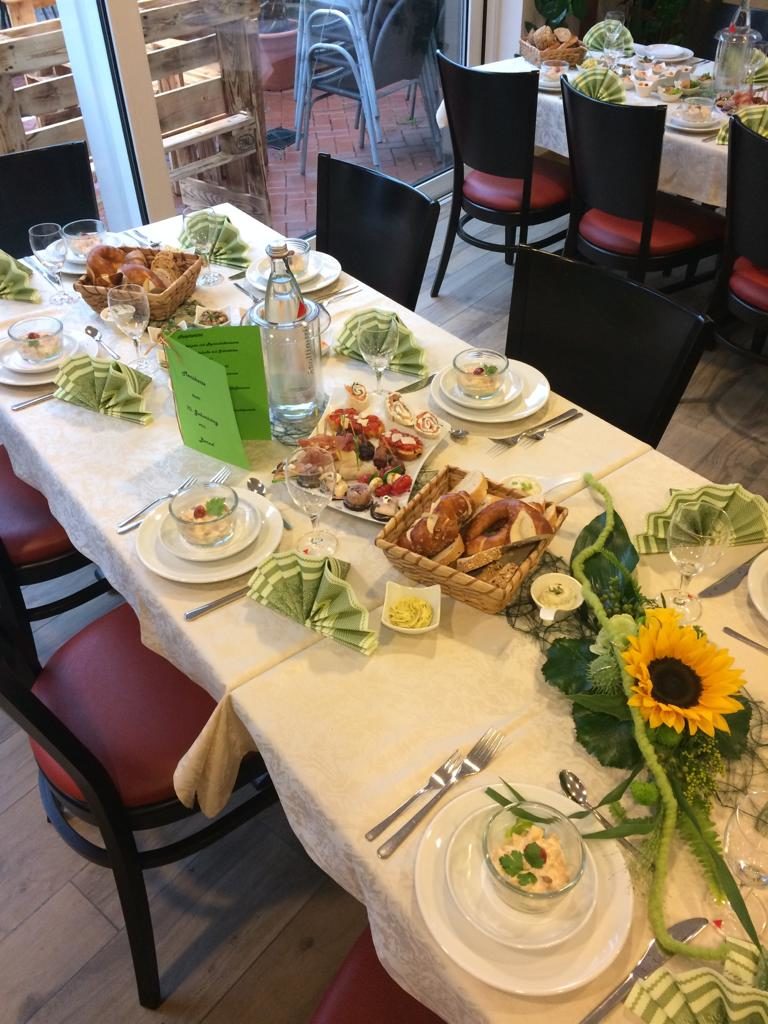 Mit Sonnenblumengesteck eingedeckter Tisch. In grün gehalten mit Weißen Tischdecken. Brezeln und Laugenbrötchen liegen in einem schön angerichten Körbchen. Daneben steht ein Teller mit kleinen Snacks. Auf den Tellern ist in einem Glasschälchen ein Salat angerichtet.